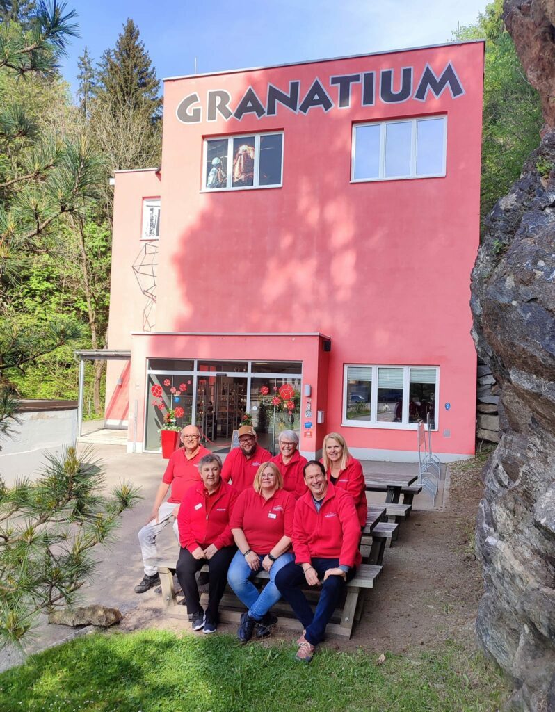 Das Team des Granatium Radenthein sitzt vor dem Granatium Haus auf einer Bank und lächelt.