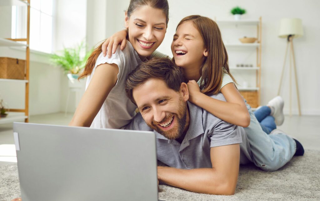 Eine Familie mit Frau, Mann und Mädchen liegen im Wohnzimmer am Teppich und schauen fröhlich in einen Laptop rein. Eine Familie mit Frau, Mann und Mädchen liegen im Wohnzimmer am Teppich und schauen fröhlich in einen Laptop rein.