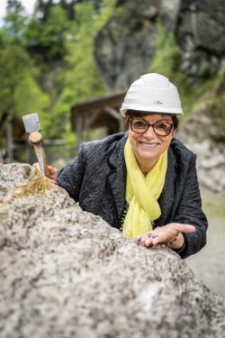 Eine Senioren steht am Schürfgelände im Granatium, hält einen Meisel in der Hand und einen Granatstein in der anderen Hand.