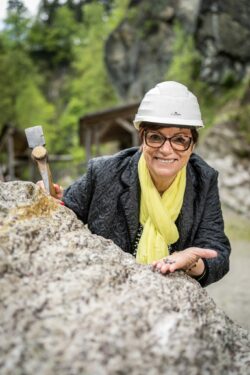 Eine Senioren steht am Schürfgelände im Granatium, hält einen Meisel in der Hand und einen Granatstein in der anderen Hand.